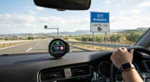 Vista desde el conductor de un coche en una autopista española (A-1 Burgos, límite 120) en 2026, con un avisador de radar legal en el salpicadero. La pantalla circular del dispositivo muestra un check verde, '118 km/h' en un límite de '120', 'SISTEMA LEGAL OK' y 'SIN INCIDENCIAS: RADARES FIJOS ACTIVOS'. Se ve una señal de tráfico de límite 120 y un pórtico de radar fijo en la distancia.